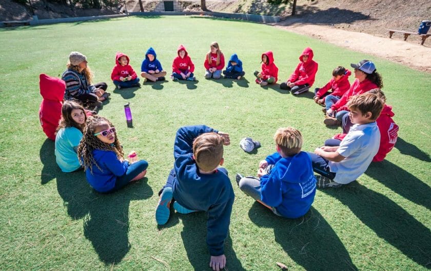 Group of pali students in blue and red sweatshirts sit in circle on grass